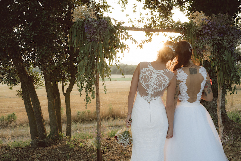 A lesbian couple in their wedding dresses holding hands looking at the sky